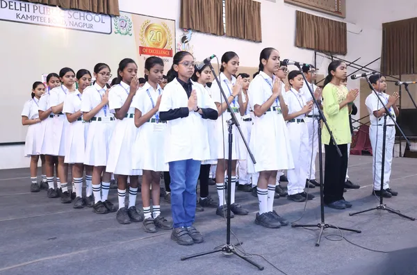 DPS (Kamptee Road) Grade VIII-B students standing on stage in school uniforms with hands folded, in front of microphones and school banner.