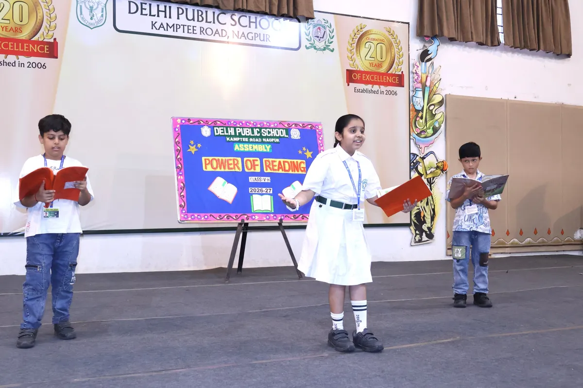 Three students holding books stand on a school stage in front of an assembly backdrop board, with a girl in white uniform at the center.