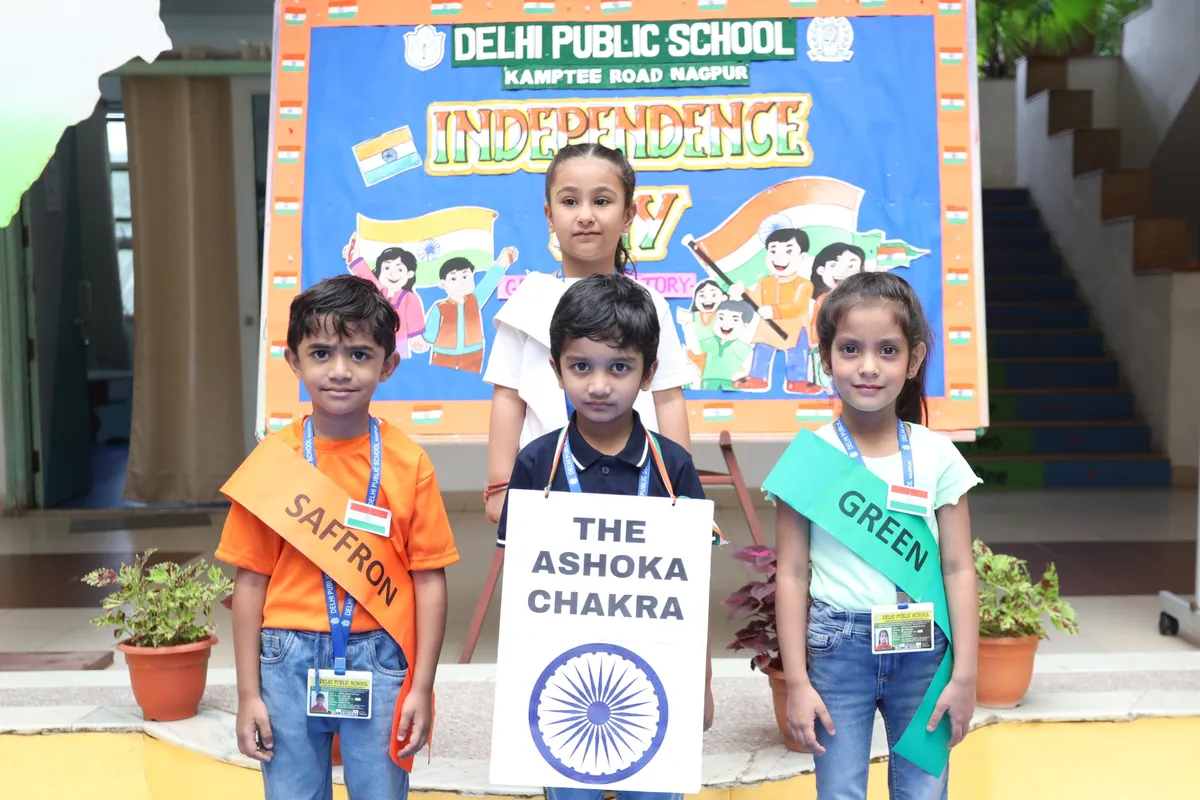 Four young students pose in front of an Independence Day board, three wearing saffron and green sashes and one holding an Ashoka Chakra sign.