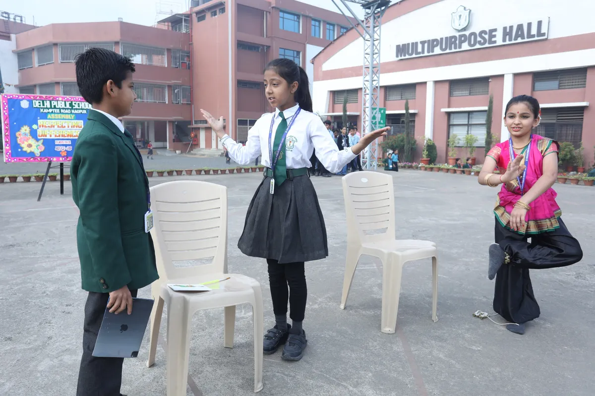 Three students perform a skit in a school courtyard, one in a green blazer, one in uniform gesturing, and one in a pink dance costume balancing on one leg, with a banner and school building behind