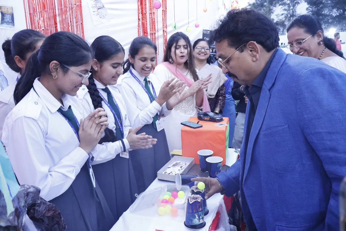 A man in a blue blazer looks at a science exhibit while schoolgirls in DPS (Kamptee Road) uniform clap beside him.