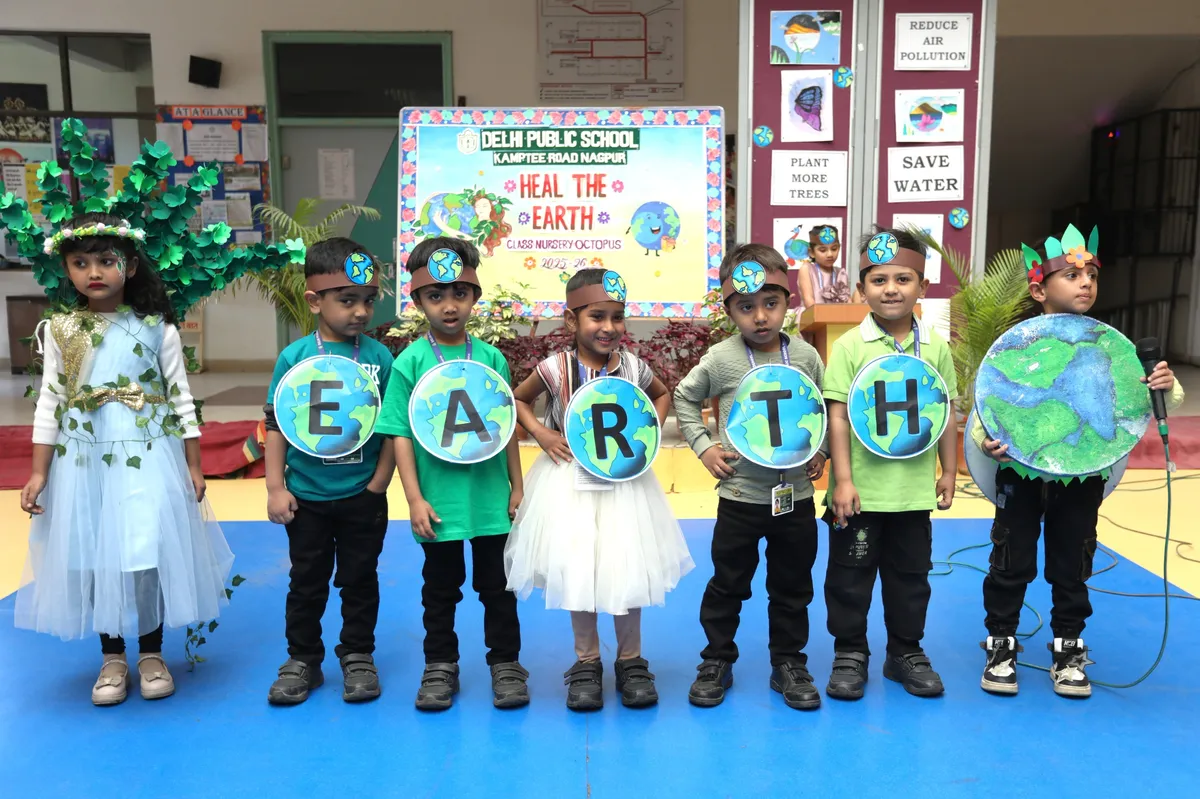 Young children in green costumes hold globe-shaped cards spelling EARTH on a blue stage, with a child in a leaf costume and another holding a large globe, in front of an environment-themed backdrop, at DPS (Kamptee Road)