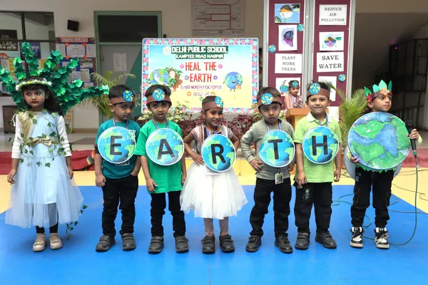 Young children in green costumes hold globe-shaped cards spelling EARTH on a blue stage, with a child in a leaf costume and another holding a large globe, in front of an environment-themed backdrop, at DPS (Kamptee Road)