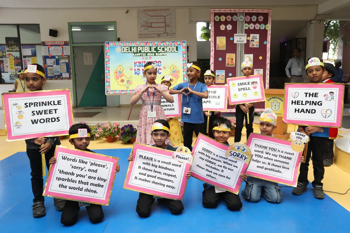 Group of young students in headbands holding pink-framed placards about kind words and manners, standing on a blue mat in front of a school banner.
