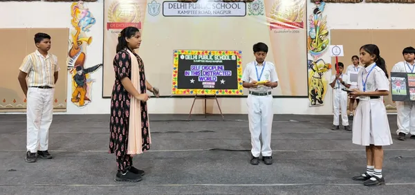 Students in school uniform and a woman in a dark patterned kurta stand on a stage in front of a board and a backdrop, apparently during a school presentation or assembly. - DPS (Kamptee Road)
