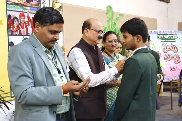 Indoor ceremony where a man in a maroon vest pins a badge on a student in a green blazer, with a teacher and a woman looking on and a decorated welcome banner behind them. - DPS (Kamptee Road)