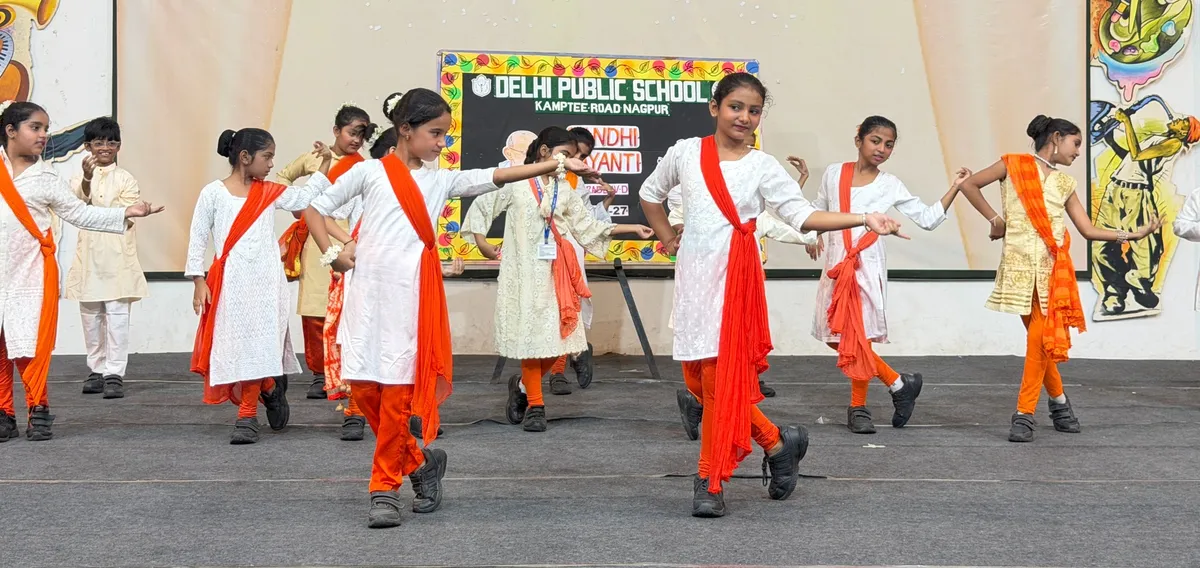 DPS (Kamptee Road) students in white outfits with orange scarves performing a group dance on a stage in front of a decorated backdrop banner