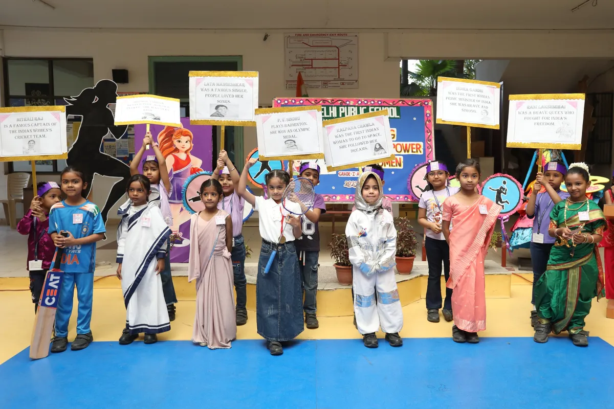 Group of young students in costumes holding placards and props such as a cricket bat and badminton racket, standing in a row in front of a decorated backdrop, during DPS (Kamptee Road) assembly.