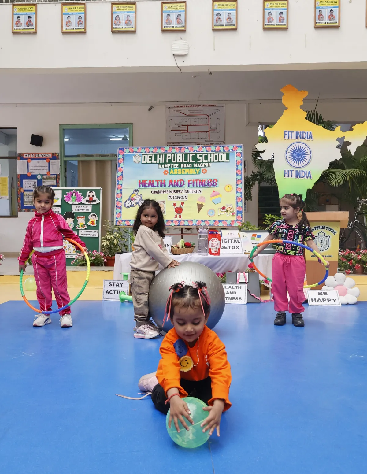 Four young children with hula hoops and exercise balls on a blue stage floor during a health and fitness assembly, with a decorated backdrop banner behind them. DPS (Kamptee Road).