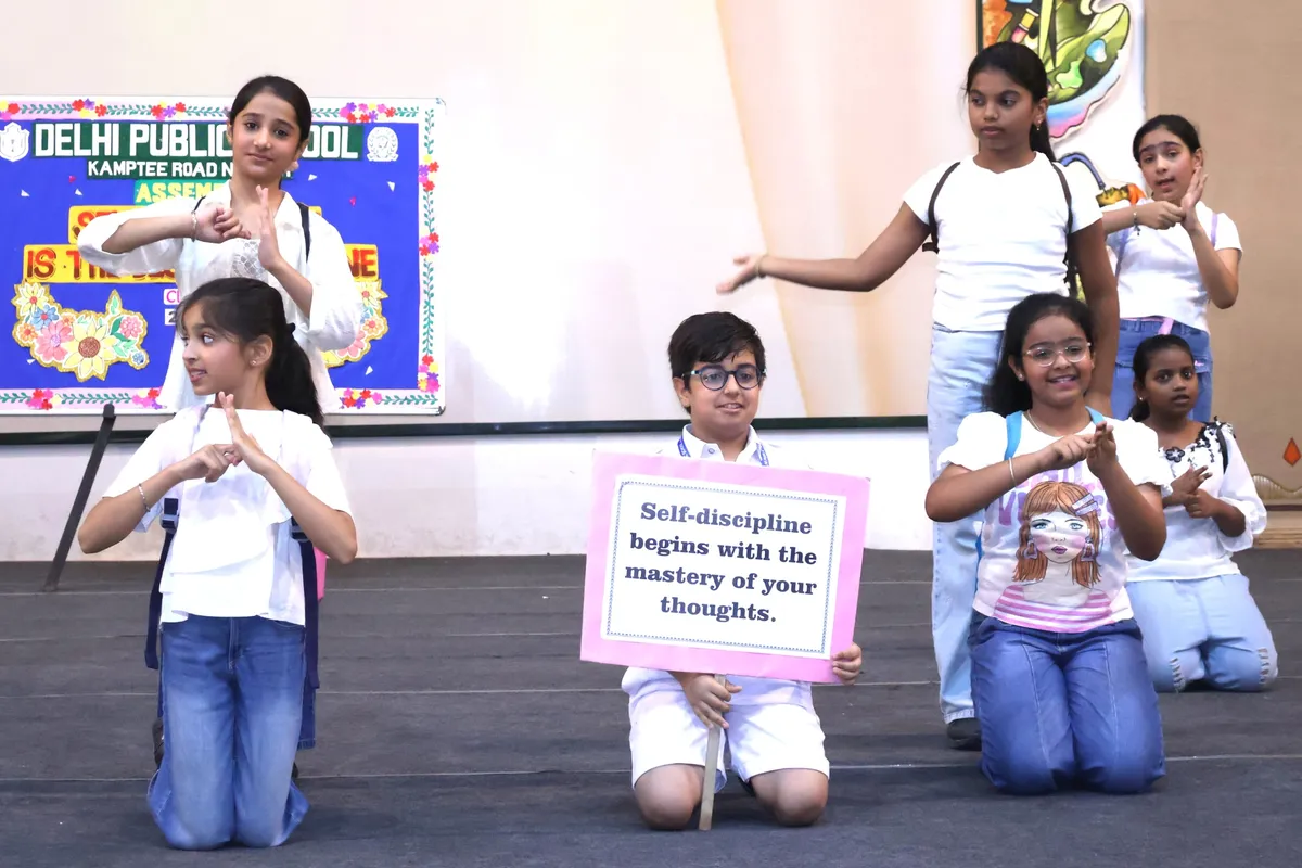 Group of schoolchildren performing a skit on a stage, one boy holding a placard, with a decorated school board behind them.