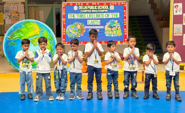Nine young children in white shirts and jeans stand in a row with palms pressed together on a blue mat in front of a decorated board and a large globe. - DPS (Kamptee Road)