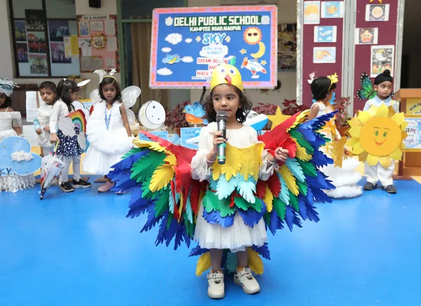 A young child in a colorful bird costume with paper wings speaks into a microphone on a blue stage, with other children in sky-themed costumes and a decorated backdrop behind. - DPS (Kamptee Road)
