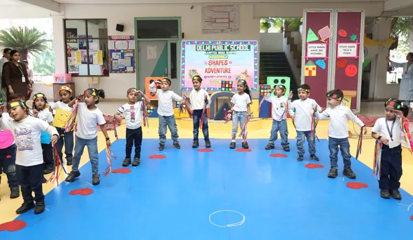 Young children in white shirts and headbands holding colorful ribbons stand in a row on a blue floor during a school assembly, with a decorated backdrop banner behind them.