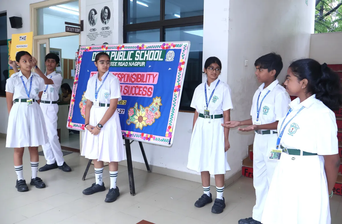 Six students in white school uniforms with ID lanyards stand and gesture around a decorated board in a school corridor.