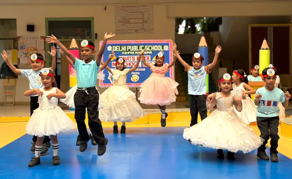 Young children in party outfits and headbands dancing with raised arms on a blue stage in front of a school banner and large pencil decorations. - DPS (Kamptee Road)