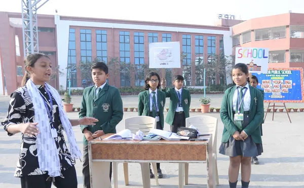 DPS (Kamptee Road) students in green blazers stand around a table with books outdoors in a school courtyard, with signs and a decorated board in the background.