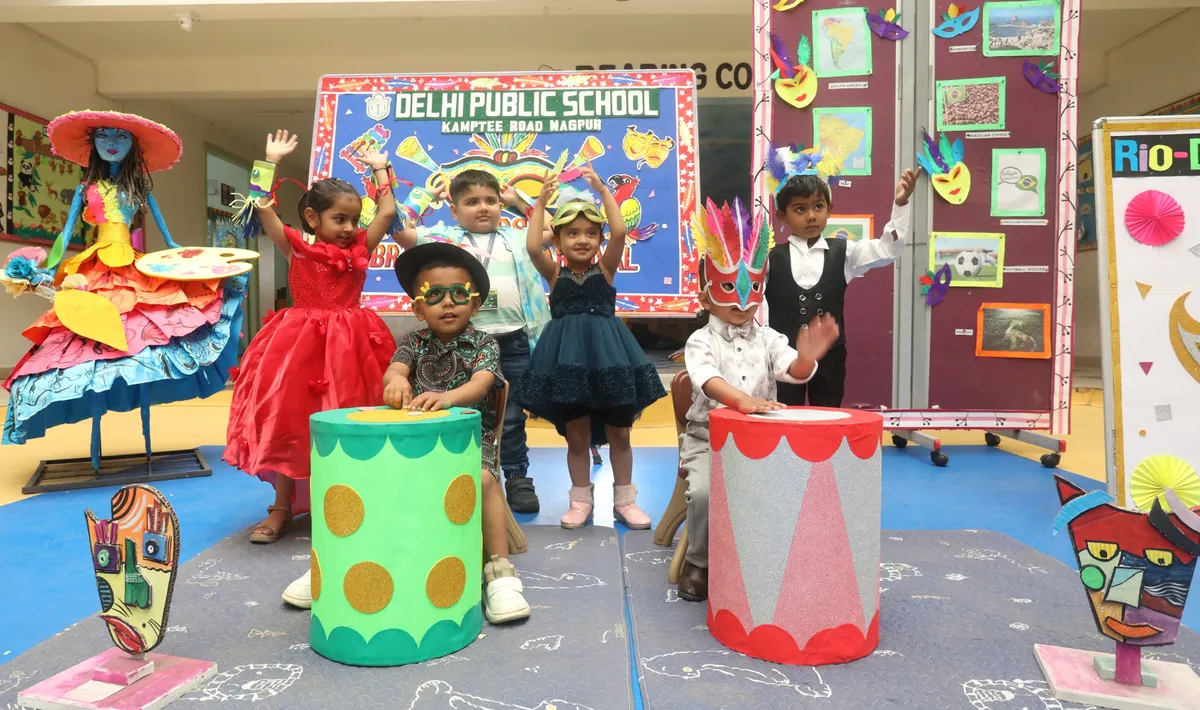 Six young children in colourful costumes and masks posing with drum props, a carnival-themed backdrop and a decorated figure on a DPS (Kamptee Road) school stage.