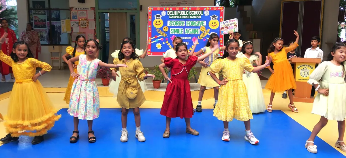 Group of young girls in colorful dresses performing a dance on a blue stage in front of a decorated board, with a podium and a few onlookers behind them. - DPS (Kamptee Road)