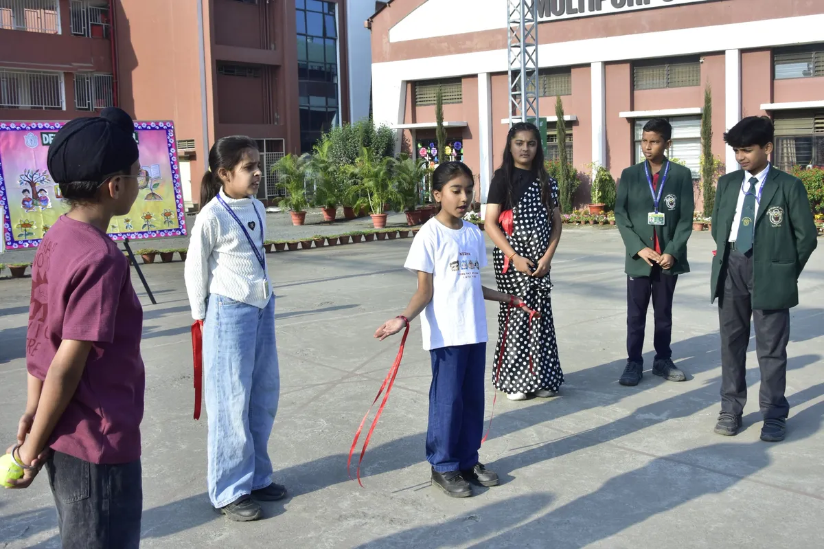 Six students stand outdoors in a school courtyard, with a girl in a white t-shirt holding red ribbons in the center and a decorated board and school buildings in the background.