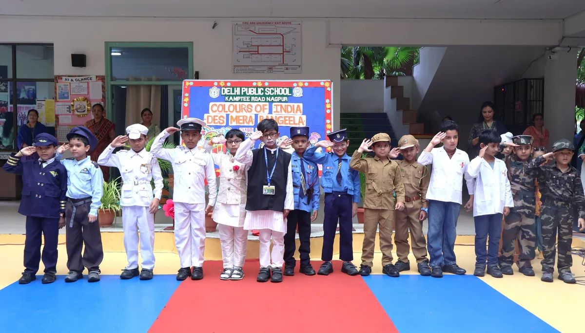 Children in uniform costumes including police, naval, army and doctor outfits saluting in a row in front of a stage backdrop banner on a colorful floor at DPS (Kamptee Road)