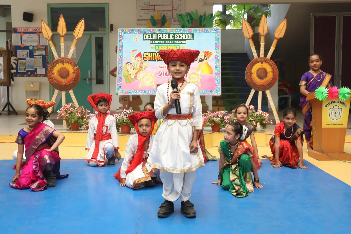 Young boy in traditional costume speaking into a microphone on a school stage, with seated children in traditional dress, decorative spears and shields, and a backdrop banner behind him.