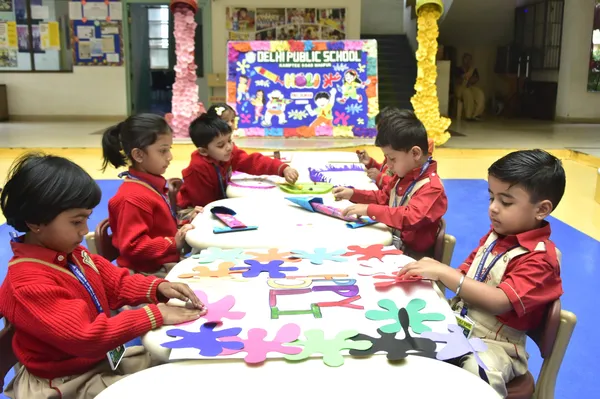 Young children in red sweaters sit around tables doing a colourful paper splash craft activity in a classroom with a decorated backdrop at DPS (Kamptee Road).