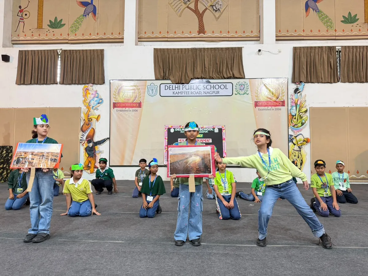 Students in green costumes perform on a hall stage at DPS (Kamptee Road), three holding up landscape pictures while others sit behind them.