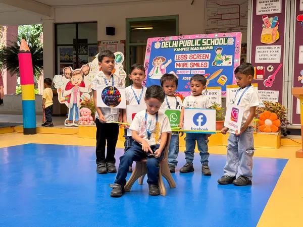Six young children holding social media app cutouts perform a skit on a blue floor in front of a DPS (Kamptee Road) banner about less screen time.