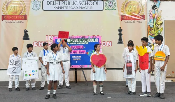 DPS (Kamptee Road) students in white uniforms on a stage holding props such as a paper heart, a cola bottle and fries beside a World Heart Day board.