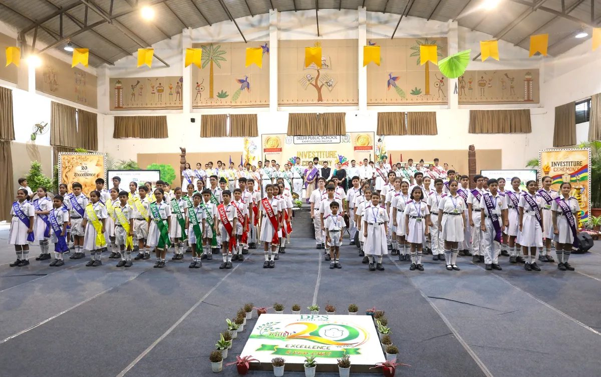 Students in white uniforms wearing colored sashes standing in a decorated school hall during Investiture Ceremony, with floral anniversary display on the floor