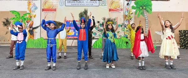 Children in colorful costumes, including blue outfits and a peacock dress, performing a dance on a stage with a decorated backdrop. - DPS (Kamptee Road)