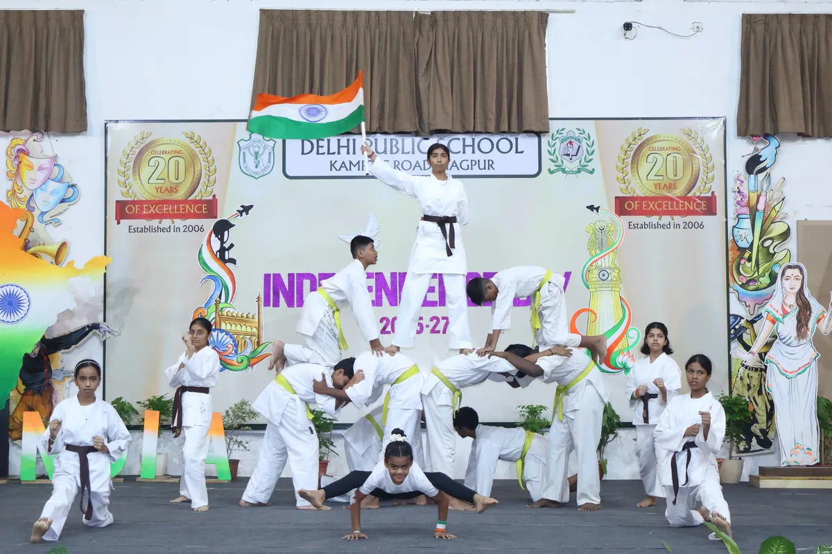 DPS (Kamptee Road) students in white martial arts uniforms form a human pyramid on a stage while one holds an Indian flag, in front of an Independence Day banner.