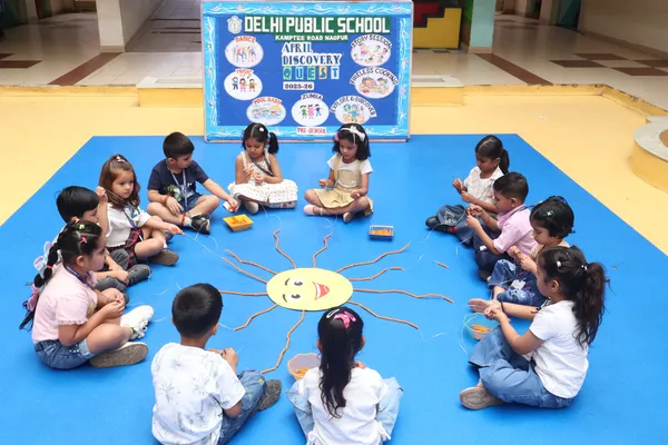 Preschool children sitting in a circle on a blue mat doing a craft activity around a paper sun, with DPS (Kamptee Road) banner in the background.