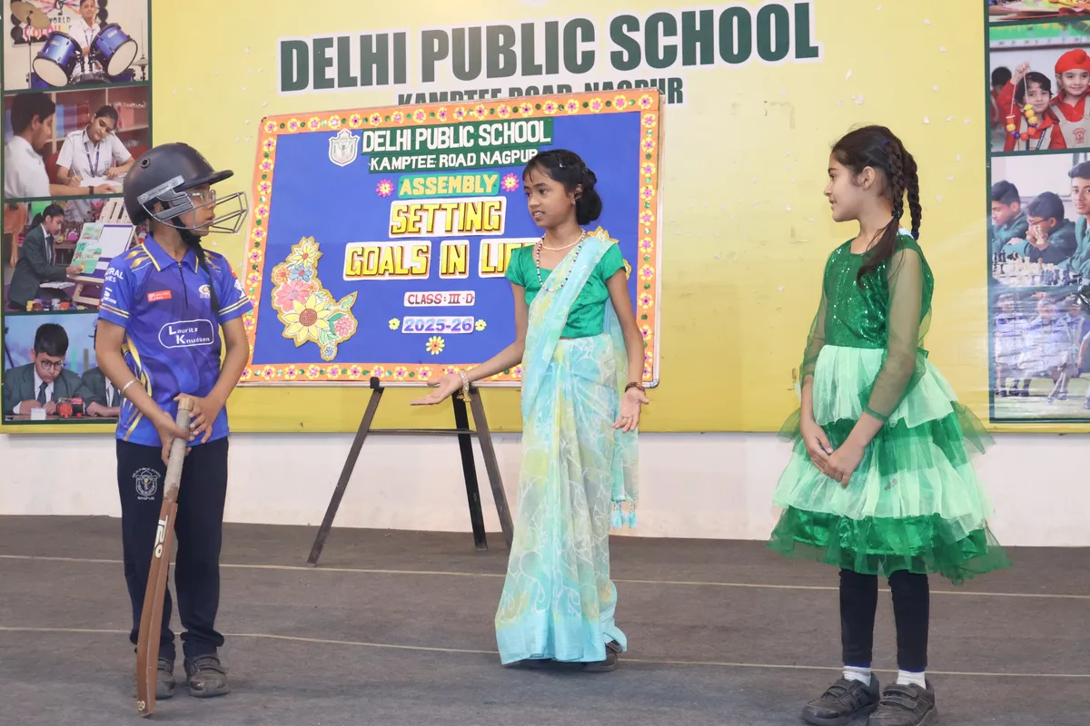 Three children in costume, one in cricket gear and helmet, perform on a stage in front of a decorated assembly backdrop board at DPS (Kamptee Road).