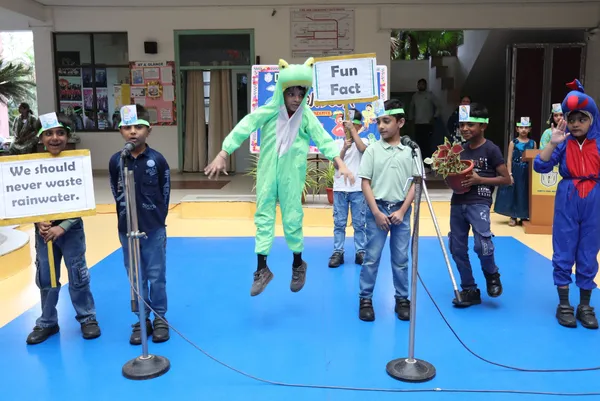 Young students on a blue stage performing a skit, one in a green frog costume jumping, others holding signs and standing at microphones