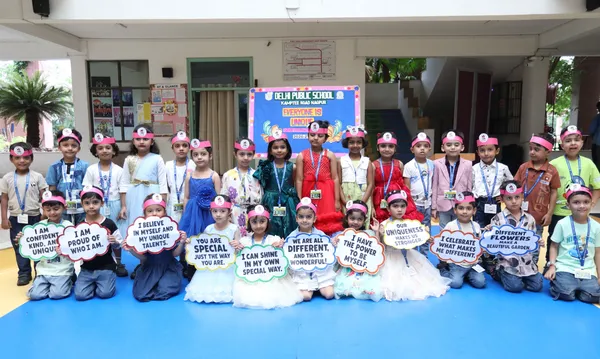 Group photo of young students in pink headbands holding speech-bubble signs with positive messages, posed in front of a DPS (Kamptee Road) school banner on a blue floor.