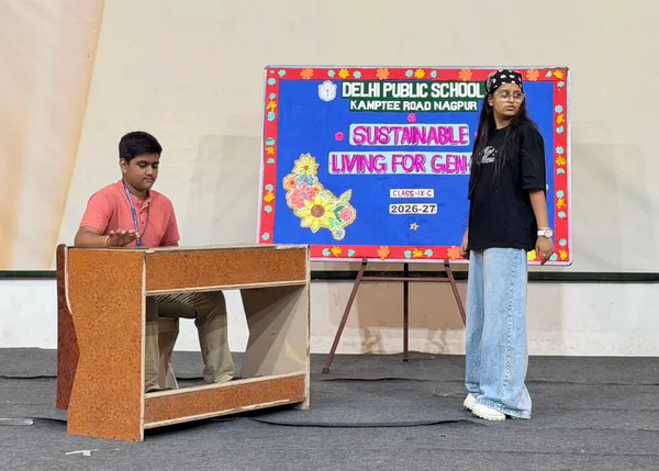 Two students performing on a stage, one seated at a wooden desk and one standing beside a decorated board on an easel. - DPS (Kamptee Road)