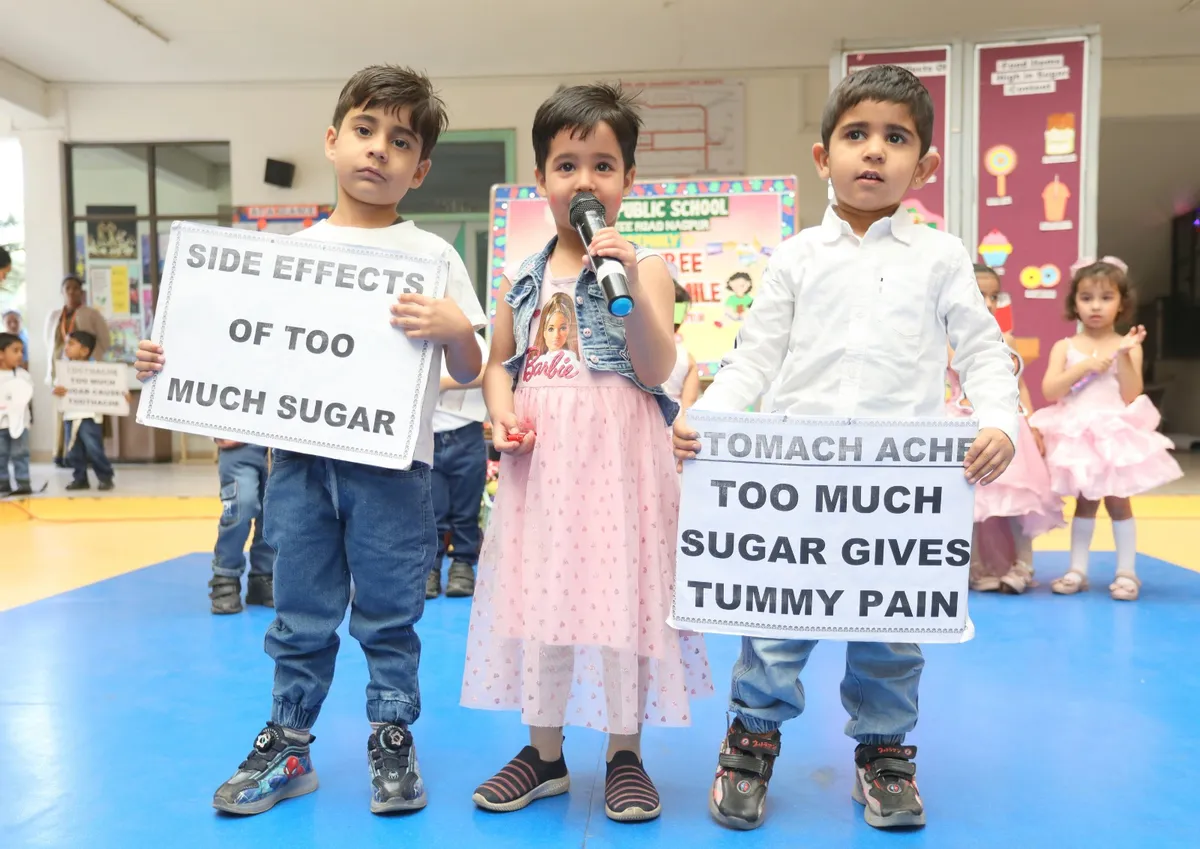 Three young children stand on a blue floor holding placards about the side effects of too much sugar, one speaking into a microphone, with other children and posters behind them. DPS (Kamptee Road).