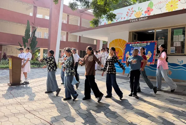 DPS (Kamptee Road) students walking in a line across a paved courtyard near a podium and a school building, with a decorated board in the background.