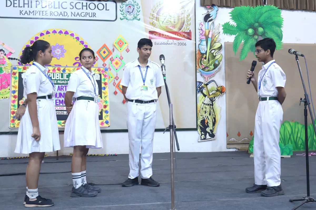 Four students in school uniforms stand on a stage with microphones in front of a decorated backdrop, one boy speaking into a microphone. - DPS (Kamptee Road)