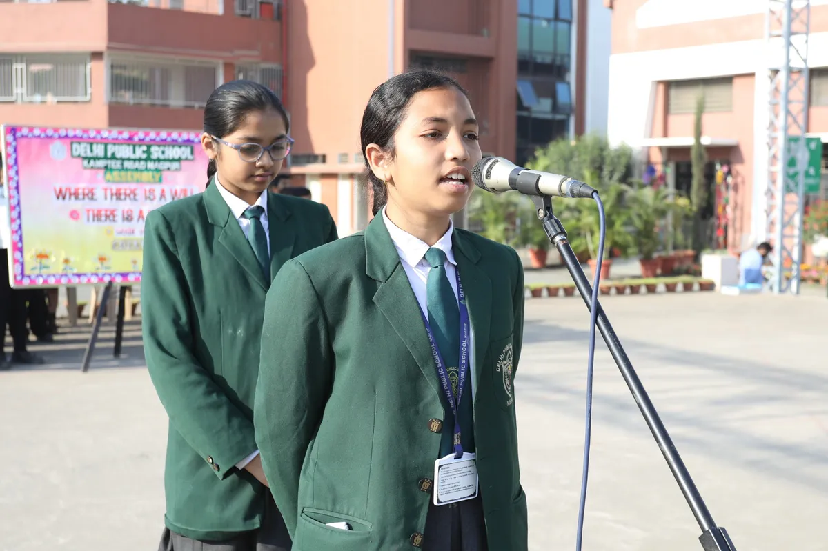 Two students in green blazers at DPS (Kamptee Road) school assembly, one speaking into a microphone, with a decorated banner behind them.
