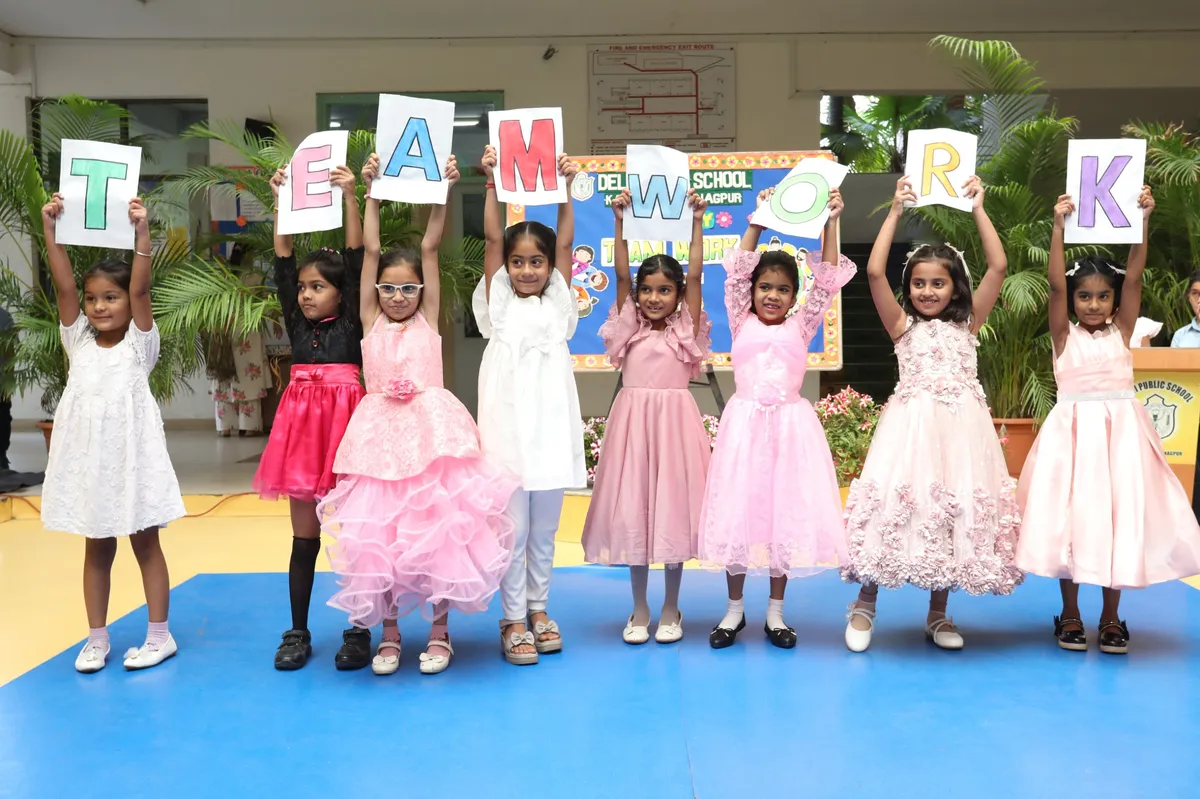 Nine young girls in party dresses stand in a row on a blue stage mat, each holding up a large letter card. DPS (Kamptee Road).