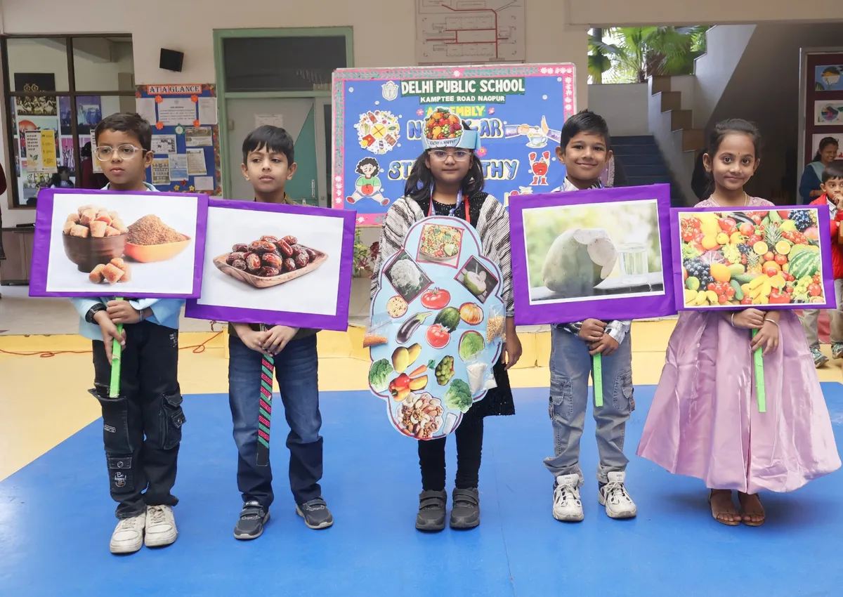 Five young students stand on a blue floor holding food picture boards of fruits, dates and nuts in front of a DPS (Kamptee Road) school assembly backdrop.