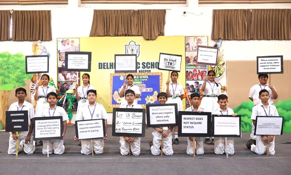 Group photo of about 16 Grade VI-D students in school uniform holding placards with messages, in front of a decorated stage backdrop at DPS (Kamptee Road)