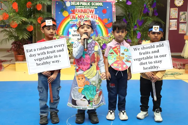Young children on a stage, one in a fruit and vegetable costume speaking into a microphone and two holding placards about eating fruits and vegetables, in front of a rainbow-themed backdrop. DPS (Kamptee Road).