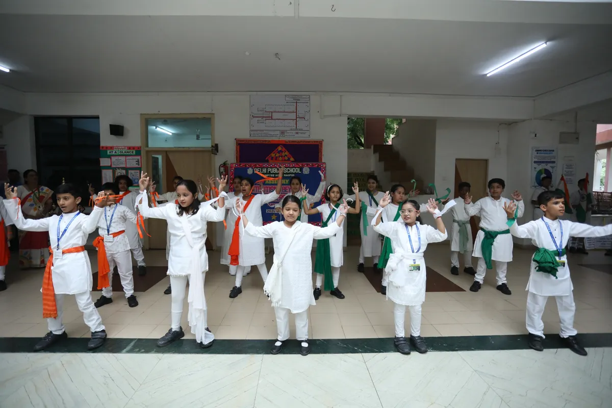 Group of schoolchildren in white outfits with orange and green sashes performing a hand-raised dance in a school hall at DPS (Kamptee Road).