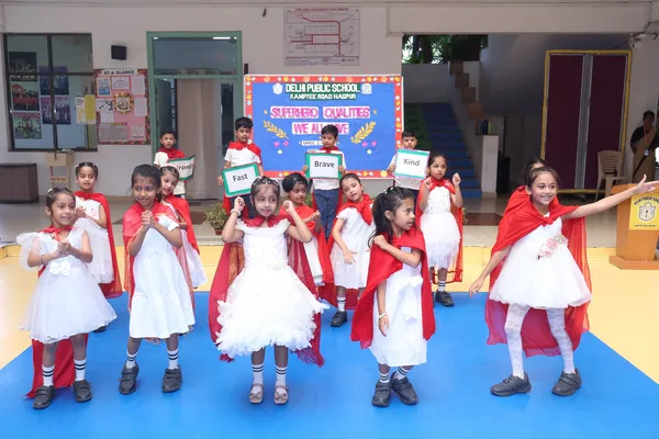 Young children in white dresses and red capes perform on a blue mat in front of a decorated board, some holding word cards. - DPS (Kamptee Road)