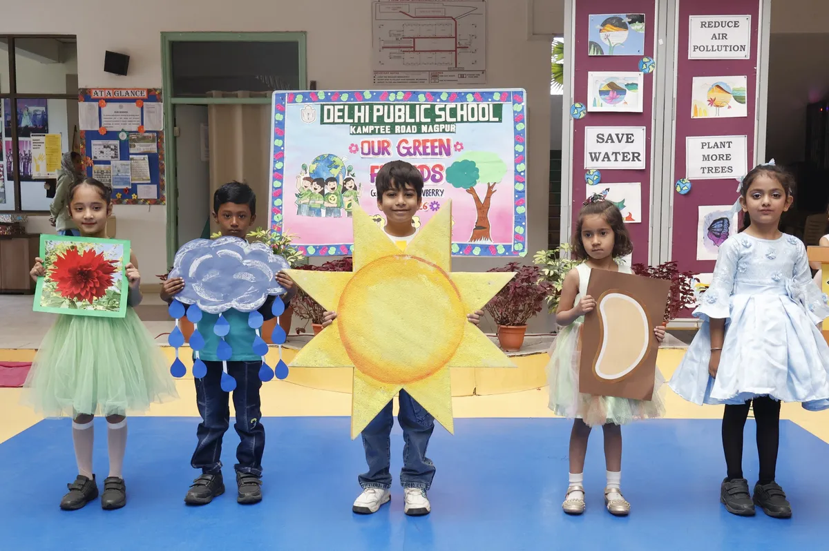 DPS (Kamptee Road) Grade I Strawberry students in costumes holding cut-outs of a flower, rain cloud, sun, and moon, standing on a blue mat in front of environmental awareness banner and display boards