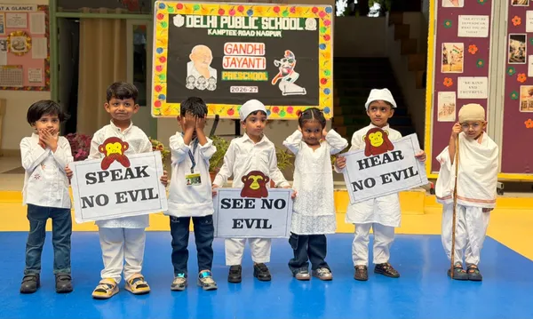 Preschool children in white costumes standing in a row on a blue floor in front of a decorated board during the Gandhi Jayanti assembly at DPS (Kamptee Road).