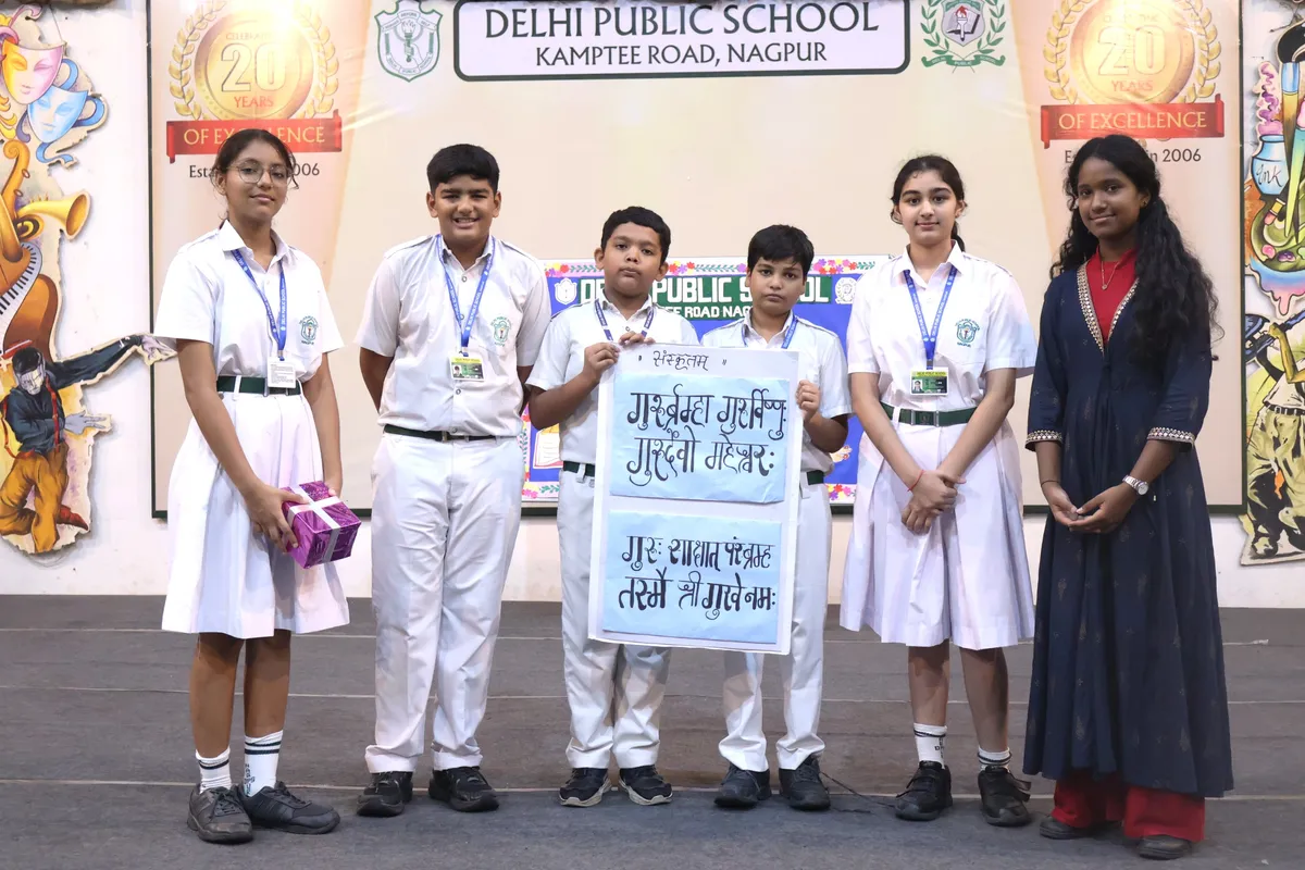 Group of five students in school uniform and a woman in dark dress on stage with placard in Devanagari and gift box, DPS (Kamptee Road) backdrop